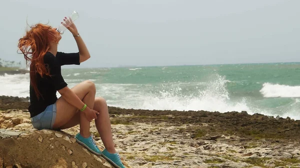 Jeune femme aux longs cheveux roux assise sur la plage et à l'eau potable en République dominicaine — Photo