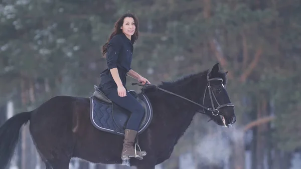 Bela mulher de cabelos compridos montando um cavalo preto através da neve na floresta — Fotografia de Stock