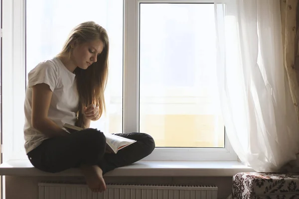 Jovem linda menina adolescente loira sentada no peitoril da janela, estudando e lendo um livro didático — Fotografia de Stock