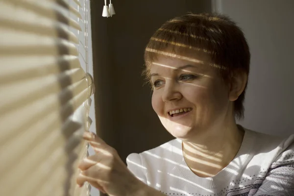 Женщина средних лет, стоящая перед окном при дневном свете, мечтающая и улыбающаяся, с оттенком жалюзи на лице — стоковое фото