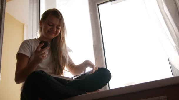Hübsche junge blonde Mädchen isst Kuchen und blättert durch das Buch sitzt auf der Fensterbank — Stockvideo