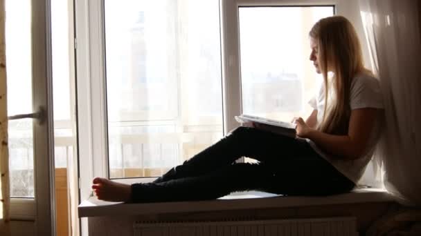 Chica rubia bastante joven hojeando el libro sentado en el alféizar de la ventana — Vídeos de Stock