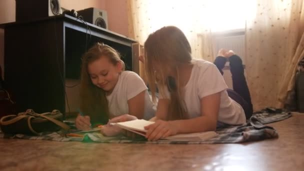 Dos adolescentes leyendo un libro y dibujando tumbados en el suelo en casa — Vídeo de stock