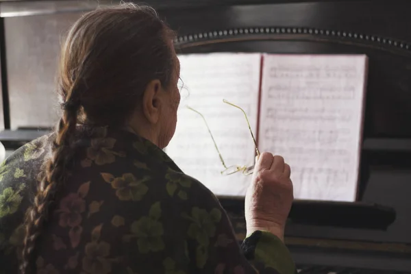 Wanita tua duduk di depan piano dan buku catatan dengan kacamata di tangannya — Stok Foto