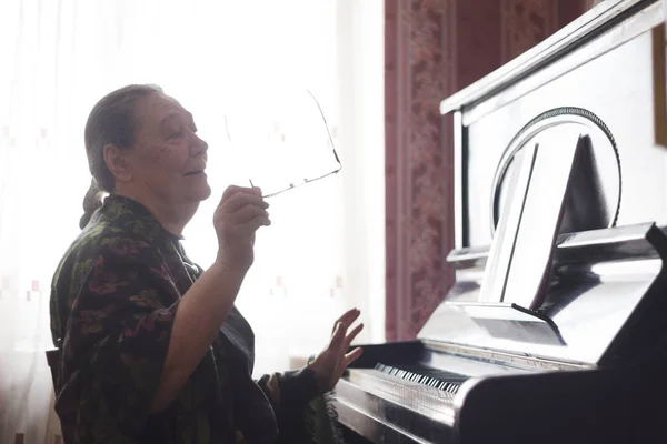 La anciana sentada frente a un piano y cantando una canción mirando un cuaderno — Foto de Stock