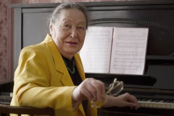 Old mempesona wanita pianis dalam jaket kuning tempat duduk di piano dengan kacamata di tangannya — Stok Foto