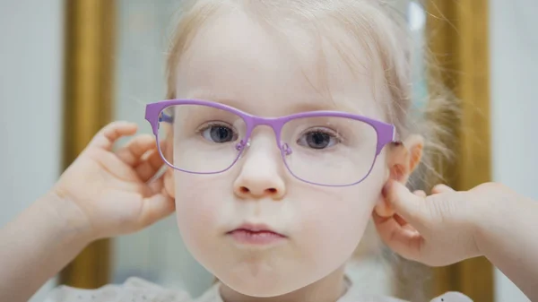 Little girl tries fashion medical glasses near mirror - shopping in ophthalmology clinic Stock Picture