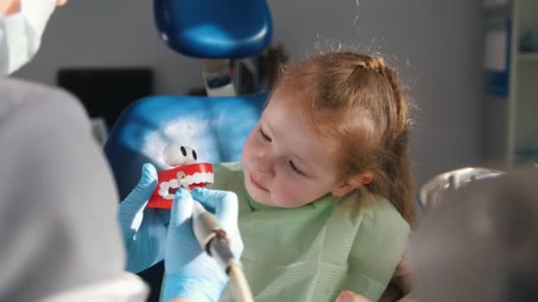En liten flicka i en stol som lyssnar på tandläkare förklaringarna om kommande förfarandet av tandborstning — Stockvideo