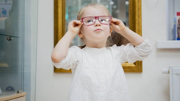 Klein meisje probeert nieuwe glazen in de buurt van spiegel - winkelen in oogheelkunde kliniek — Stockfoto
