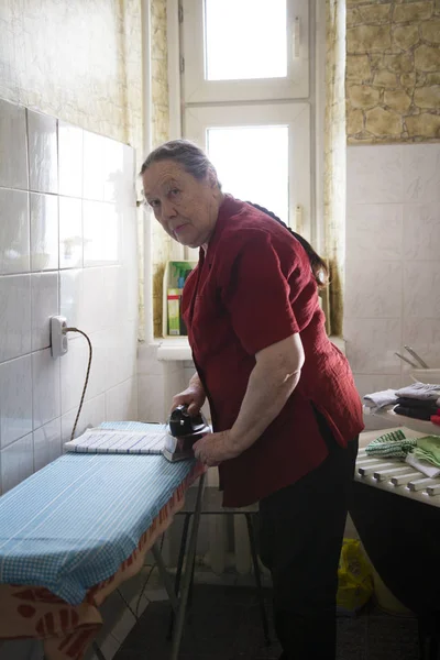 Velha senhora na blusa vermelha engomando uma toalha em casa — Fotografia de Stock