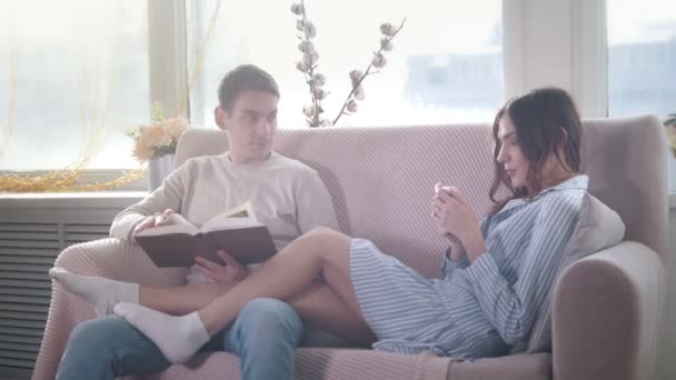 Glückliches junges attraktives Paar, das zusammen auf der Couch sitzt, der Kerl liest ein Buch, das Mädchen telefoniert — Stockvideo