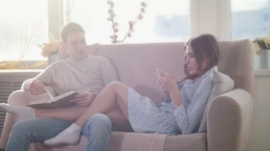 Mutlu birlikte kanepede oturan genç çekici çift adam bir kitap okur, kız kullanmak telefon