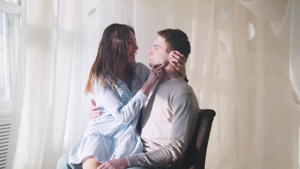 Молодий хлопець і дівчина сидять біля вікна в яскраво освітленій кімнаті, обіймаючи і цілуючи — стокове фото