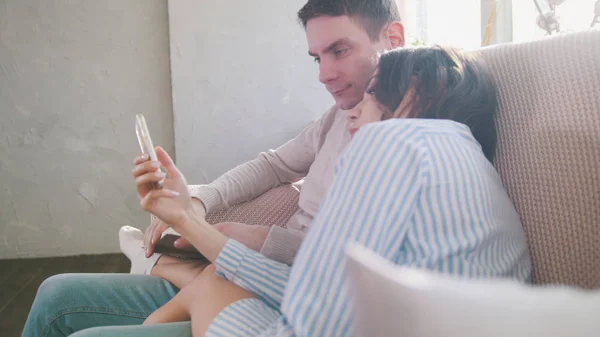 Загорелая молодая пара сидит на диване, девушка показывает что-то парню на смартфоне — стоковое фото