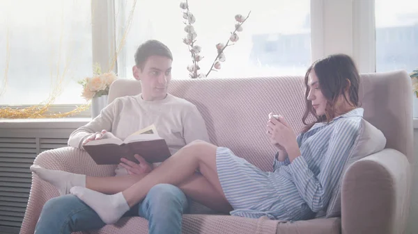 Szczęśliwa młoda atrakcyjna para siedzieli na kanapie, facet czyta książkę, Dziewczyna używać telefonu — Zdjęcie stockowe