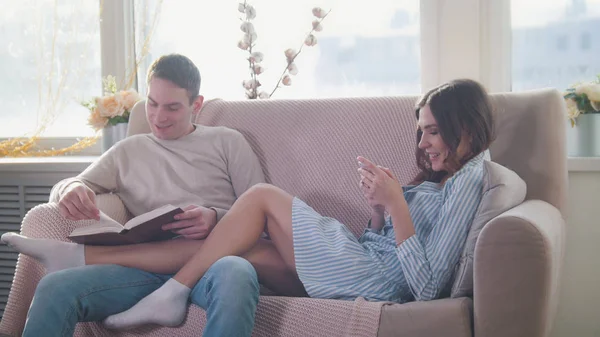 Szczęśliwa młoda atrakcyjna para siedzieli na kanapie, facet czyta książkę, Dziewczyna używać telefonu — Zdjęcie stockowe