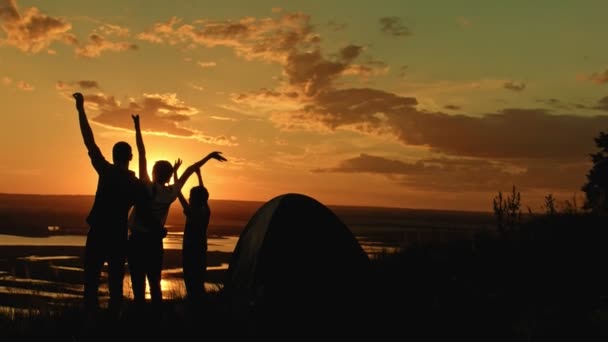 Familia joven - padre, madre e hijo en el camping - se encuentra en la colina alta al atardecer de verano y tienen feliz, silueta — Vídeos de Stock