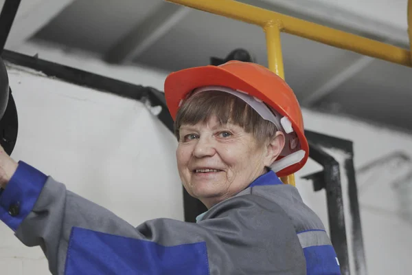 De oudere vrouw in het werkoverall en rode helm op de productiesite — Stockfoto