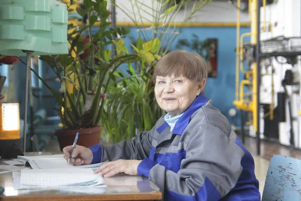Пожилая женщина в рабочей одежде пишет на рабочем месте — стоковое фото