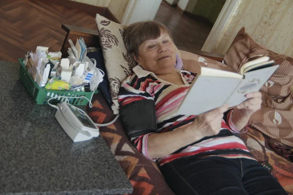 Mulher idosa doente lendo na cama com comprimidos e dispositivo para medir a pressão — Fotografia de Stock