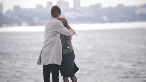 Hombre joven guapo calienta a la chica con su abrigo en el puente en un día ventoso — Vídeos de Stock