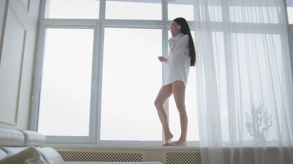 Schöne junge Frau in weißem Herrenhemd und Socken, die auf der Fensterbank spaziert, lacht und telefoniert glücklich — Stockfoto
