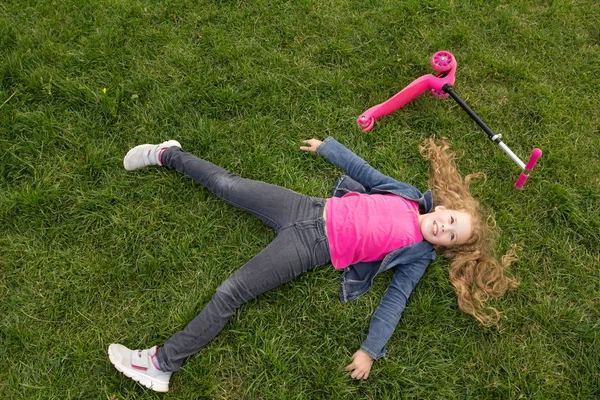 ピンクのスクーターで緑の草の上に横たわるジーンズのジャケットを着て笑顔の子女の子 — ストック写真