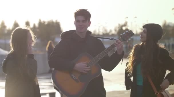 Dwie dziewczyny uśmiechnięty facet gra na gitarze na zachód słońca na zewnątrz — Wideo stockowe