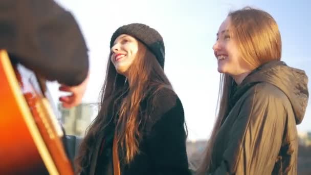 Счастливые девушки поют с парнем, играющим на гитаре на закате — стоковое видео