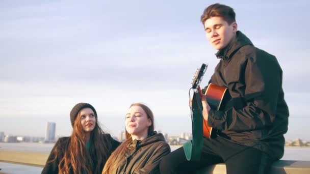 Een paar gelukkige vrienden zingen met de gitaar bij de zonsondergang buitenshuis — Stockvideo