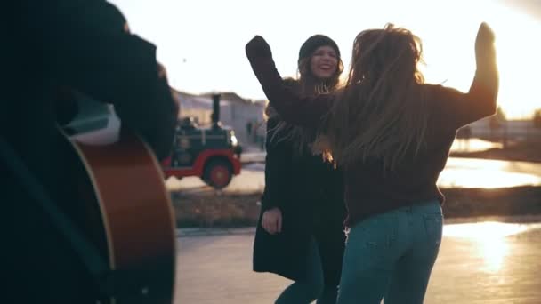 Guitarrista frente a chicas felices con el pelo largo saltando al atardecer — Vídeo de stock