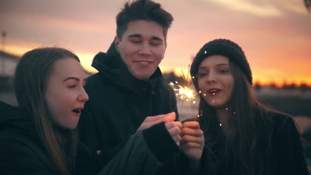 Drie gelukkige vrienden met plezier samen met sparkler bij zonsondergang buitenshuis — Stockvideo