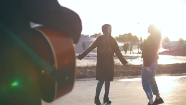 Homem tocando guitarra na frente de jovens dançando meninas ao pôr do sol — Vídeo de Stock