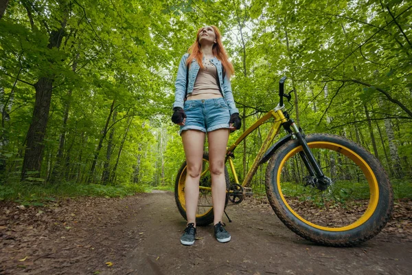 Стройная молодая женщина, стоящая в зеленом лесу на жирном велосипеде — стоковое фото