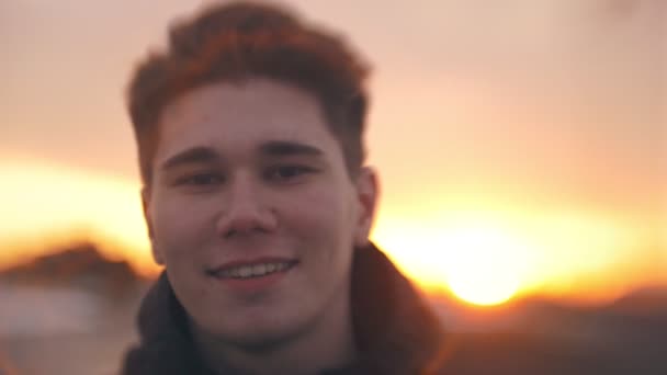 Портрет молодого улыбающегося мужчины, смотрящего в камеру на закате — стоковое видео