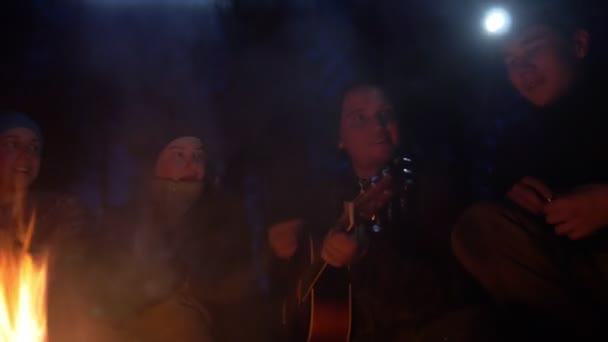 Jóvenes amigos felices en la hoguera en el bosque nocturno cantando canciones y tocando la guitarra — Vídeo de stock