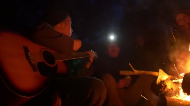 Jóvenes amigos junto a la hoguera en la madera nocturna cantando canciones y tocando la guitarra — Vídeo de stock