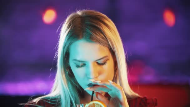 Πανέμορφη ξανθιά νεαρή γυναίκα κάθεται δίπλα στο περίπτερο μπάρμαν - πίνοντας ένα ποτό από το άχυρο και κοιτάζοντας στην κάμερα - νέον μπλε φωτισμός — Αρχείο Βίντεο