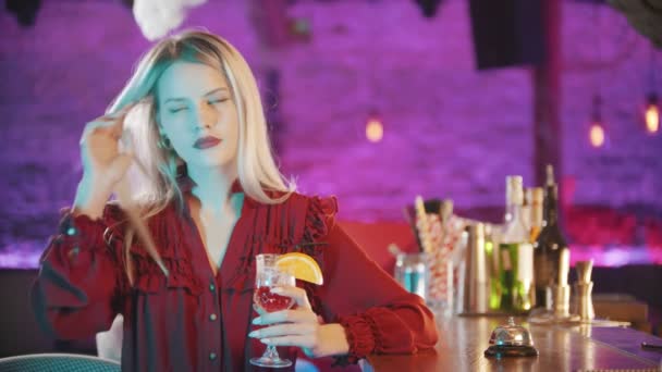 Gyönyörű szőke fiatal nő piros rúzzsal ül a pultos állvány és játszik a hajával, és kezében egy ital - neon kék világítás — Stock videók