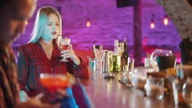 Barmen standının yanında oturan güzel sarışın genç kadın. Pipetten içecek içen adam. Ön planda telefonuyla oturan adam.