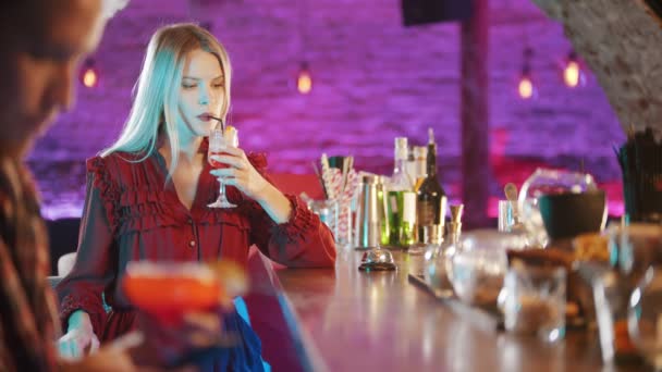 Gyönyörű szőke fiatal nő ül a pultos standnál - szalmaszálból iszik - egy férfi ül a telefonjában az előtérben és iszik — Stock videók