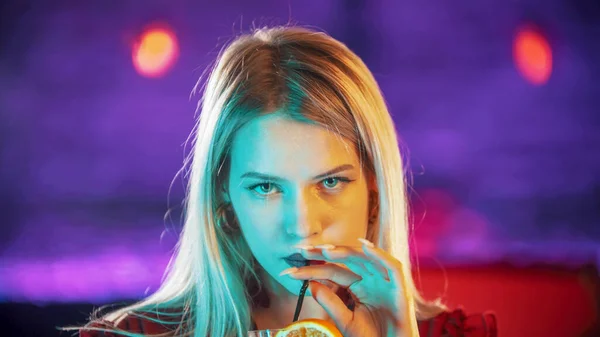 Underbar blond ung kvinna sitter vid bartendern stå - dricka en dryck från sugröret och titta i kameran - neon blå belysning — Stockfoto