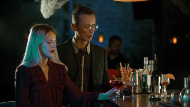 Красивая пара разговаривает и пьет коктейли у стойки в темном баре — стоковое видео