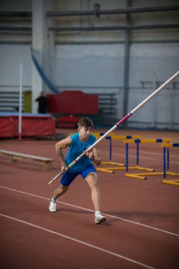 Kapalı alanda sırıkla atlayan genç bir adam podyumda elinde bir sopayla koşuyor.