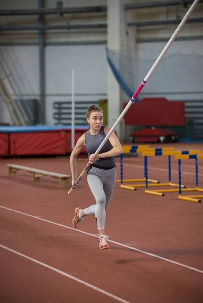 Bóveda de poste en el interior: una joven deportista corriendo por la pista con un palo en las manos —  Fotos de Stock