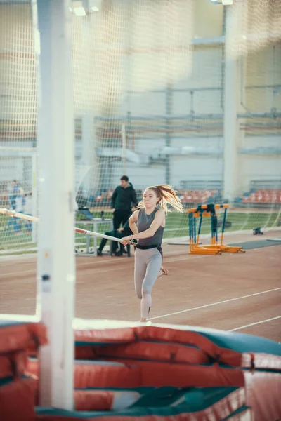 Pole bóveda en el interior - joven mujer deportiva con cola de caballo corriendo con un palo en las manos - luz brillante en el fondo —  Fotos de Stock