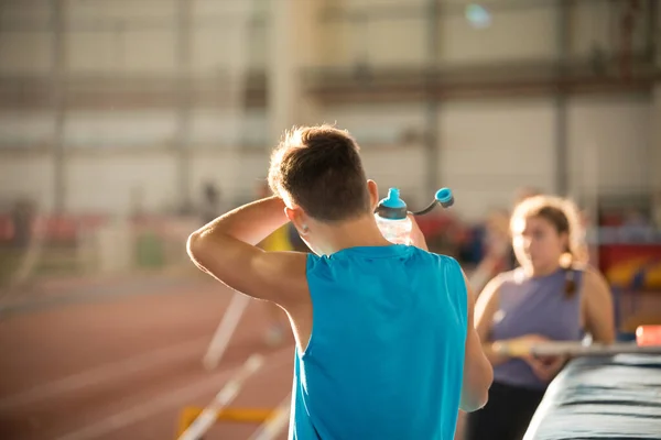 Hombre joven con camisa azul de pie en el estadio deportivo sosteniendo una botella de agua - limpiando el sudor de su frente —  Fotos de Stock