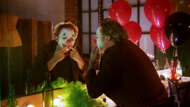 En clown sträcker ut sina läppar i ett leende och skrattar åt sig själv i spegeln — Stockvideo