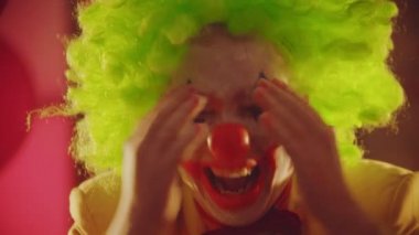 Deli bir palyaço elleriyle yüzünü kapatıyor ve ürpertici duygular ve hareketler gösteriyor.