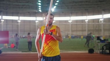 Kapalı alanda sırıkla atlayan sarı gömlekli bir adam pistte sırıkla dikiliyor derin nefes alıyor ve koşmaya başlıyor.